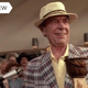 A man in a tan, brimmed hat and plaid jacket holds his first-prize chili cook-off trophy as he stands before a crowd and a microphone.